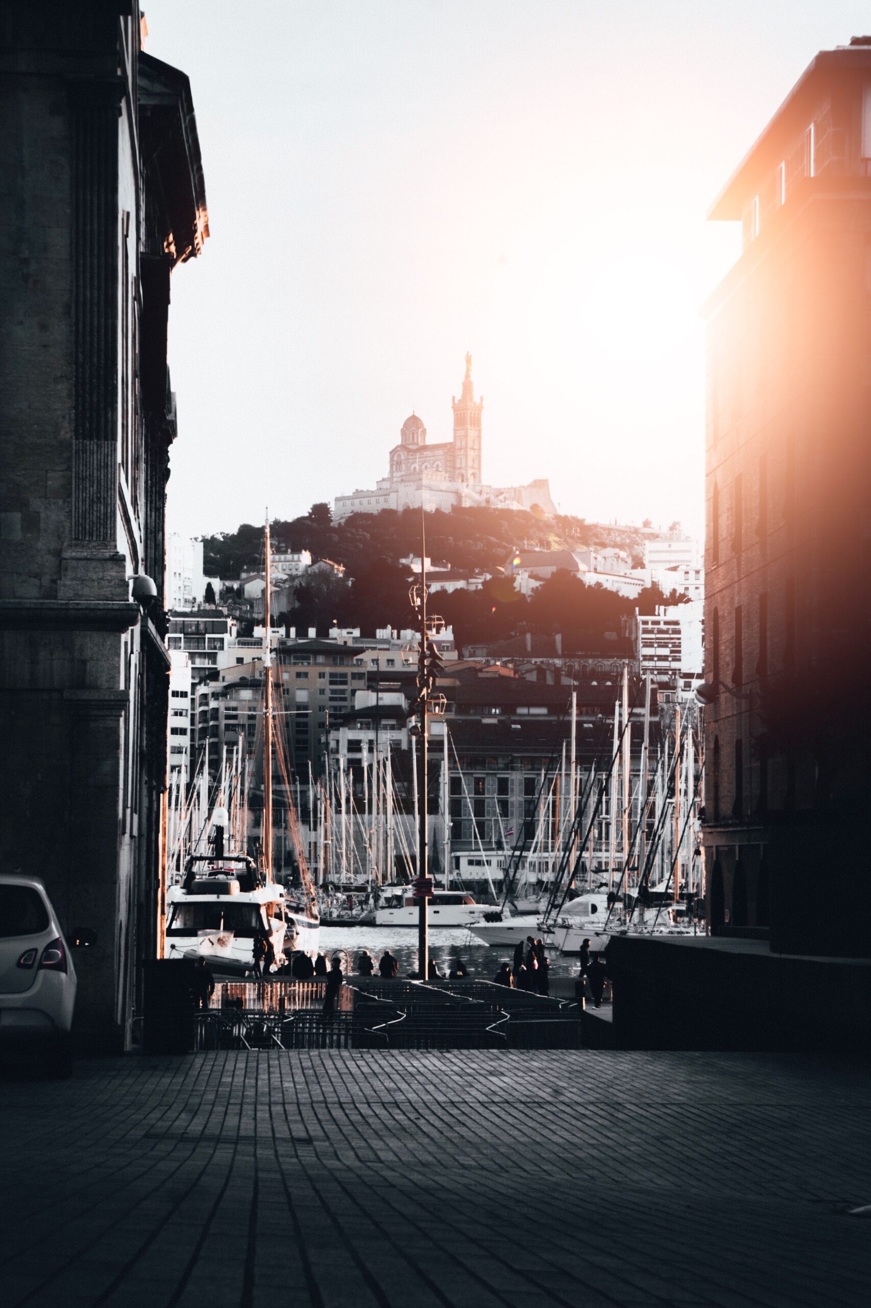 A beautiful shot of a port with lots of docked boats in the water in Marseille, France with a shining sun in the sky and a castle in the background