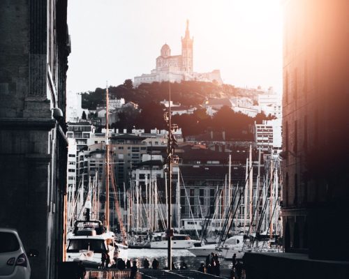 A beautiful shot of a port with lots of docked boats in the water in Marseille, France with a shining sun in the sky and a castle in the background